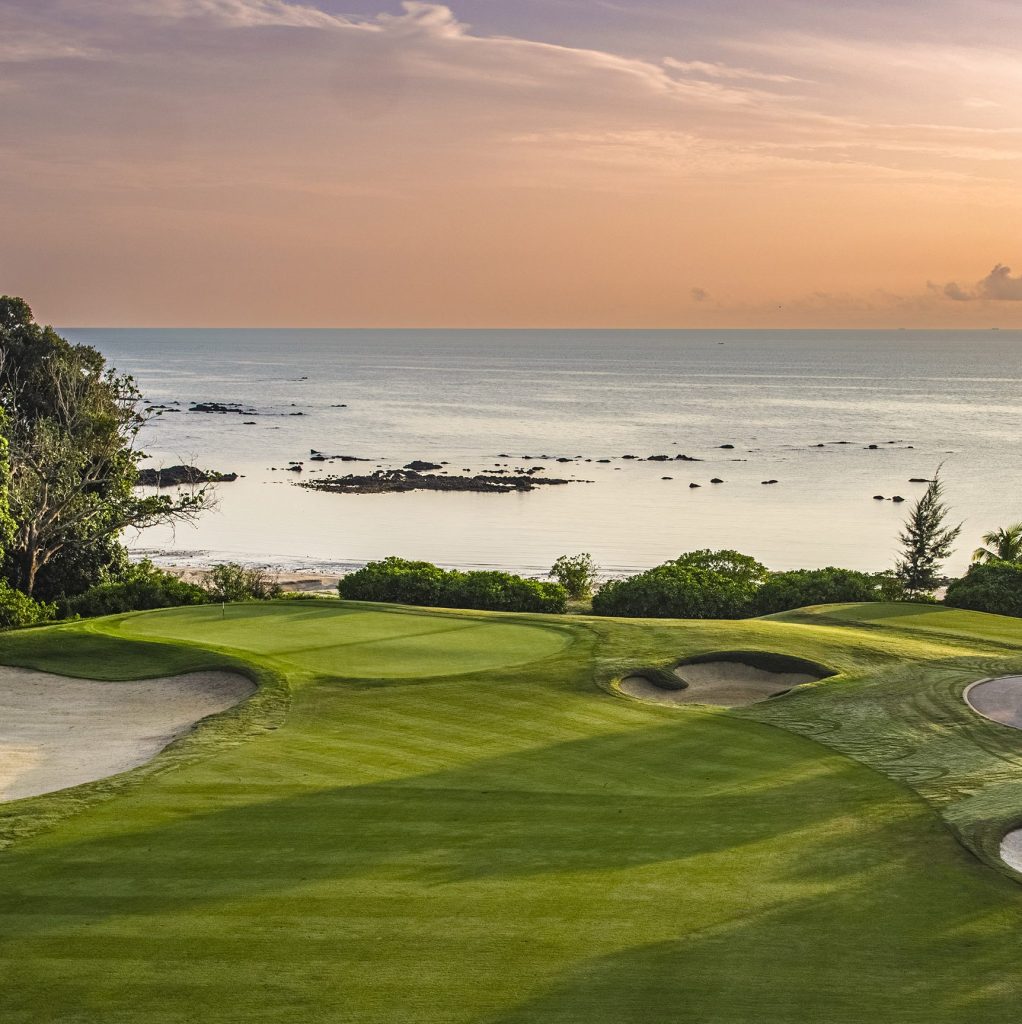 An expansive golf course with lush green fairways, neatly manicured bunkers, and towering trees lining the periphery, providing a serene backdrop for a leisurely day of golfing. The course is situated beside the South China Sea, offering breathtaking views of the sparkling blue waters and enhancing the overall tranquil atmosphere.
